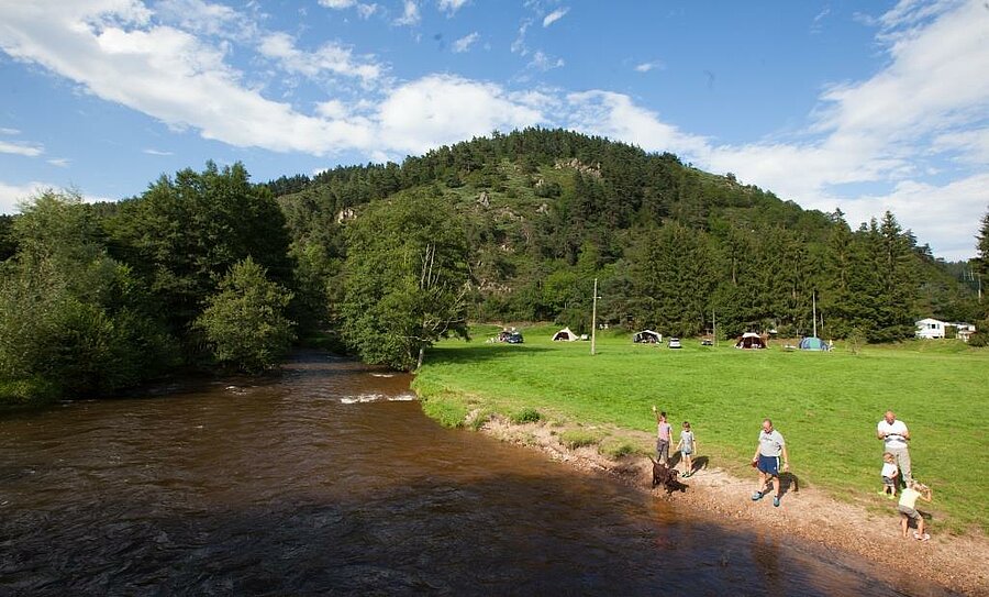 Famille se promenant au bord d’une rivière près d’un camping bord de lac, emplacements verts et forêt en arrière-plan.