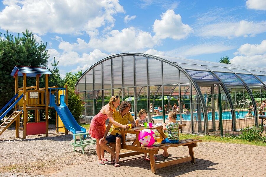 Famille profitant d’un moment convivial près de la piscine couverte et de l’aire de jeux d’un camping Sites et Paysages, lors d’un week-end de l’Ascension ensoleillé.
